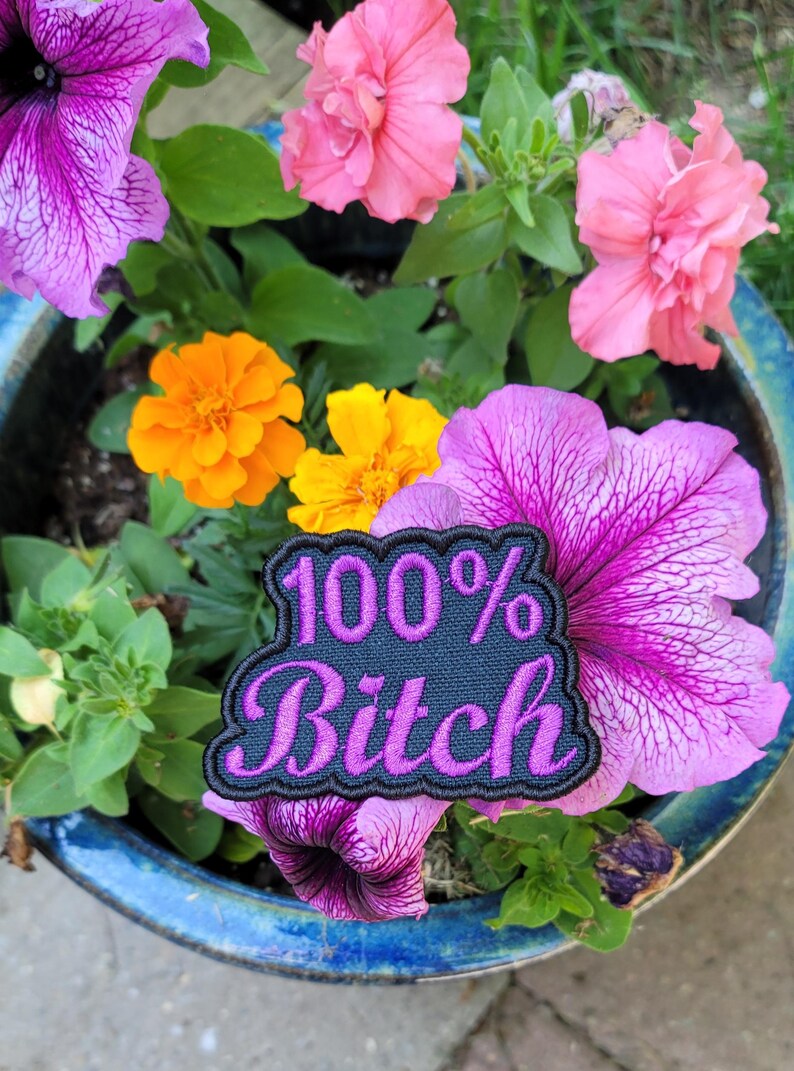 May include: A patch with the text "100% Bitch" in pink lettering on a navy blue background, surrounded by a black border. The patch is placed among vibrant pink and purple petunias, orange and yellow marigolds, and green foliage in a blue ceramic pot.