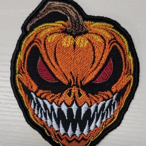May include: Embroidered patch featuring a menacing jack-o'-lantern face. The pumpkin is orange with a brown stem, red eyes, and a wide, toothy grin. The patch has a black border and is likely for a jacket or bag.
