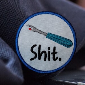 May include: A white round patch with a blue border. The patch has an embroidered image of a blue and gray seam ripper with a red tip and the word "shit" in black lettering.