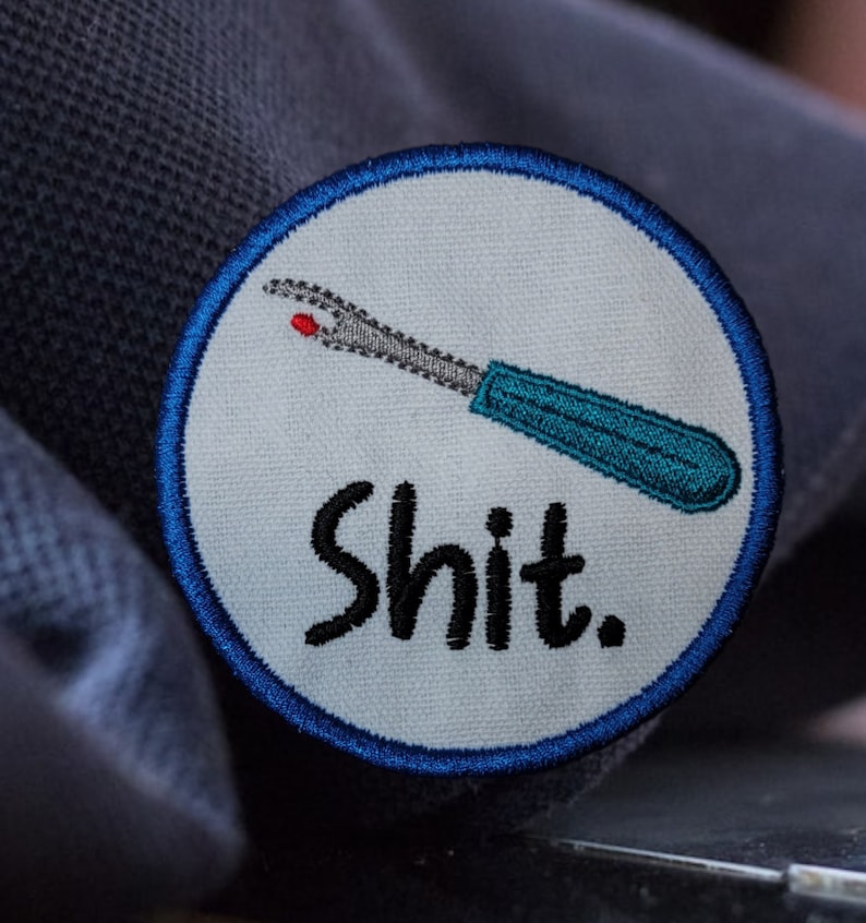 May include: A white round patch with a blue border. The patch has an embroidered image of a blue and gray seam ripper with a red tip and the word "shit" in black lettering.