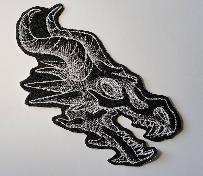 Peut inclure: Patch brod&eacute; blanc en forme de cr&acirc;ne de dragon sur un fond de tissu noir. Le cr&acirc;ne de dragon est d&eacute;taill&eacute; avec des dents ac&eacute;r&eacute;es et une expression f&eacute;roce.