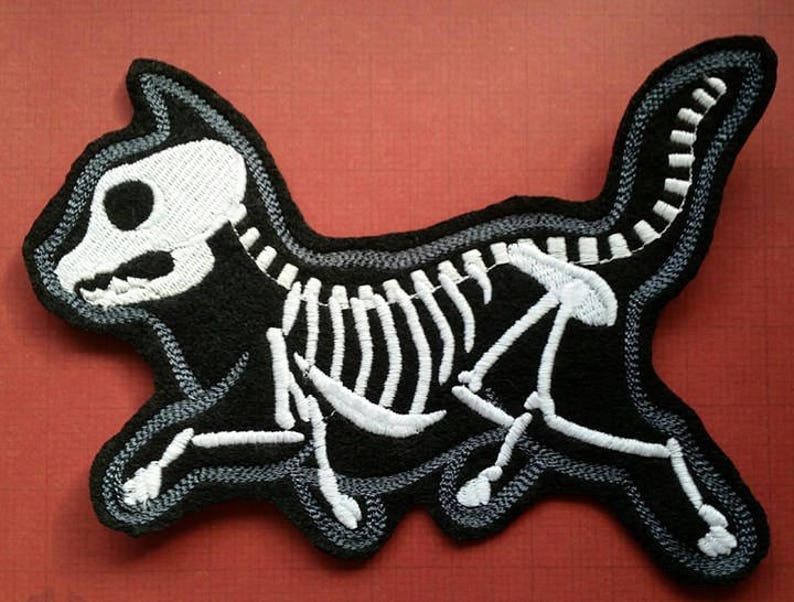 K&ouml;nnte beinhalten: Schwarz-wei&szlig; gestickter Aufn&auml;her eines Katzenskeletts. Die Katze rennt mit erhobenem Schwanz. Das Skelett ist wei&szlig; und der K&ouml;rper der Katze ist schwarz.