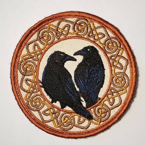 May include: Embroidered patch featuring two black ravens facing each other. The ravens are set against a cream-colored background with a brown border and gold Celtic knotwork design. The patch is round.