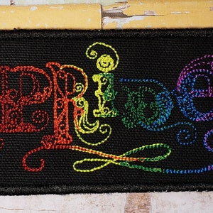 May include: A black embroidered patch with the word "Pride" in rainbow colors. The letters are stylized with swirls and flourishes.