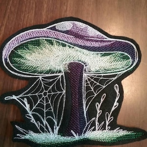 May include: Embroidered patch of a purple and green mushroom with a white spider web and green grass on a black background.