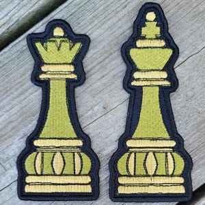 May include: Two embroidered chess pieces: a queen and a king. The pieces are lime green with gold accents and black borders. The queen has a crown, and the king has a rounded top. The pieces are on a wooden surface.