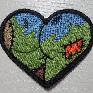 May include: A green, blue, and brown heart-shaped patch with stitched details. The patch has a black border and is made of fabric.