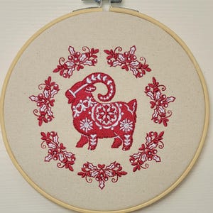 May include: A red and white embroidered goat design on a beige fabric, set within a wooden embroidery hoop. The goat is surrounded by a wreath of red holly and berries. The embroidery hoop is approximately 8 inches in diameter.