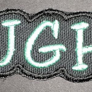 May include: A black fabric patch with a white outline and green stitching. The patch says "UGH" in a stylized font.