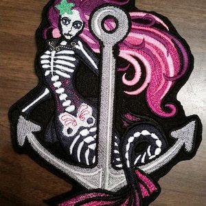 May include: An embroidered patch featuring a skeleton mermaid with pink hair, holding onto a silver anchor. The mermaid's tail is also pink and the anchor has a black background.