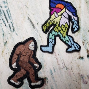 May include: Two embroidered patches featuring Bigfoot. One patch is brown and the other is a colorful mountain scene with a sunset.