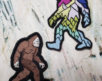 May include: Two embroidered patches featuring Bigfoot. One patch is brown and the other is a colorful mountain scene with a sunset.