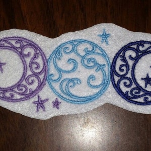 May include: Embroidered celestial design featuring a crescent moon and stars in purple, blue, and navy blue. The design is on a white, cloud-shaped felt base. The moons have intricate swirl patterns.