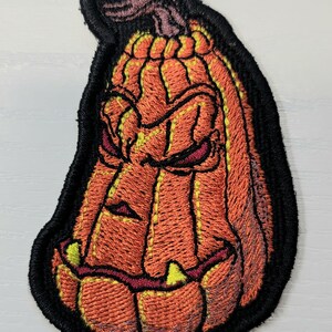 K&ouml;nnte beinhalten: Ein gestickter Aufn&auml;her in Form eines l&auml;nglichen, orangefarbenen K&uuml;rbisses mit einem d&uuml;steren, bedrohlichen Gesichtsausdruck. Der K&uuml;rbis hat tiefe vertikale Rillen, einen braunen Stiel und spitze, gelbliche Z&auml;hne, eingefasst von einem schwarzen Rand.