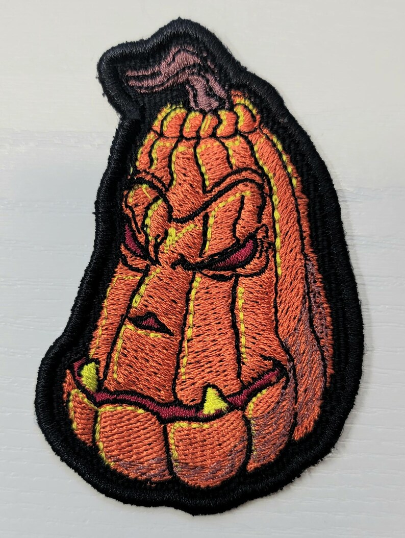 K&ouml;nnte beinhalten: Ein gestickter Aufn&auml;her in Form eines l&auml;nglichen, orangefarbenen K&uuml;rbisses mit einem d&uuml;steren, bedrohlichen Gesichtsausdruck. Der K&uuml;rbis hat tiefe vertikale Rillen, einen braunen Stiel und spitze, gelbliche Z&auml;hne, eingefasst von einem schwarzen Rand.