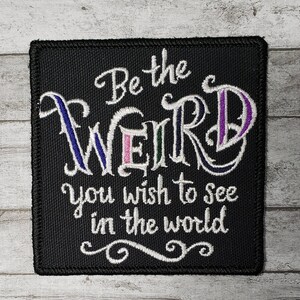 Op de afbeelding: Zwarte geborduurde patch met witte tekst die "Be the weird you wish to see in the world" luidt.