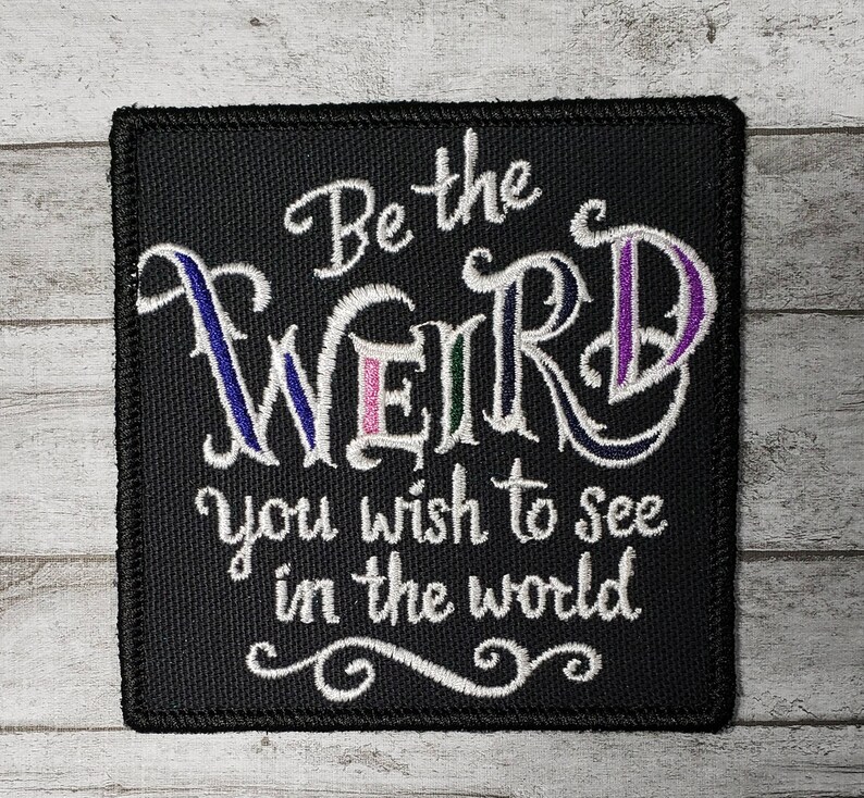 Op de afbeelding: Zwarte geborduurde patch met witte tekst die "Be the weird you wish to see in the world" luidt.