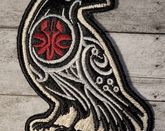 May include: Black and white embroidered patch of a raven with a red circle and design in the centre. The raven is outlined in white and has a white eye.