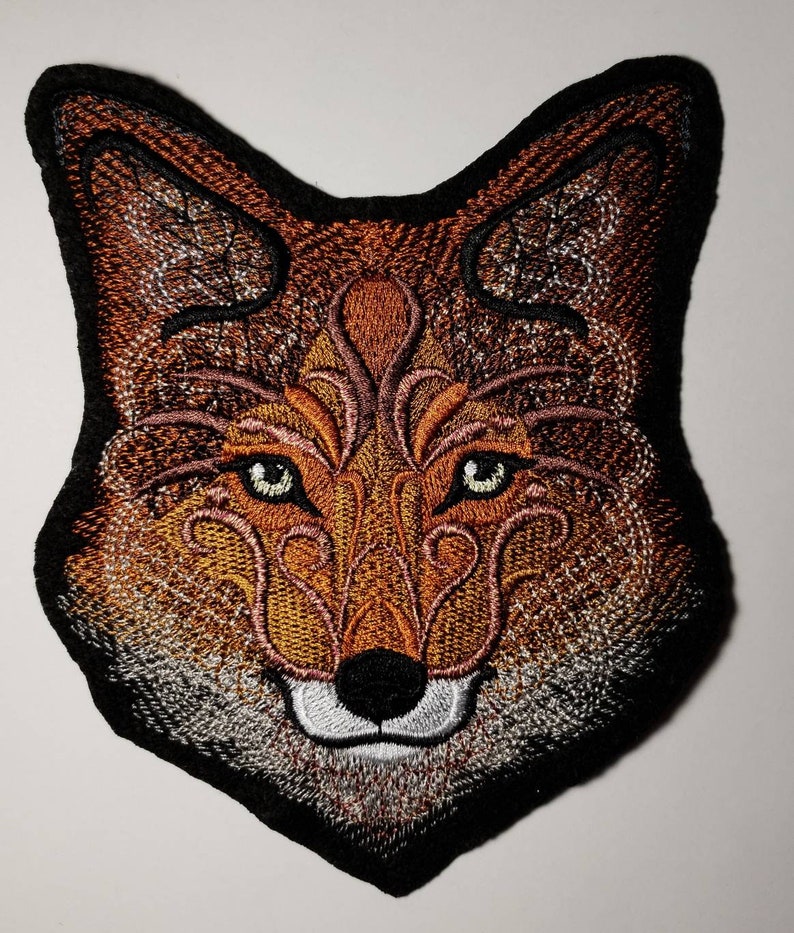 Peut inclure: Patch brod&eacute; repr&eacute;sentant la t&ecirc;te d'un renard avec des d&eacute;tails complexes dans des tons de brun, orange et blanc. Les yeux du renard sont verts et le fond est noir.