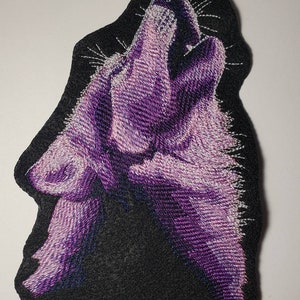 May include: Embroidered patch featuring a purple wolf howling against a black background. The wolf is rendered with detailed stitching, showcasing shades of purple and white. The patch has a black border and is likely designed for application on clothing or accessories.