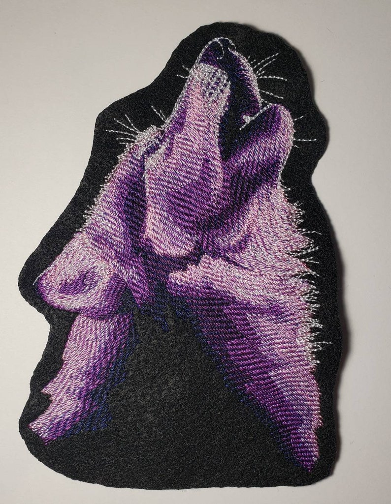 May include: Embroidered patch featuring a purple wolf howling against a black background. The wolf is rendered with detailed stitching, showcasing shades of purple and white. The patch has a black border and is likely designed for application on clothing or accessories.