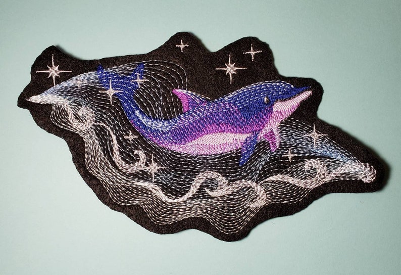 May include: Embroidered patch of a purple and blue dolphin jumping through a swirling white cloud with stars on a black background.