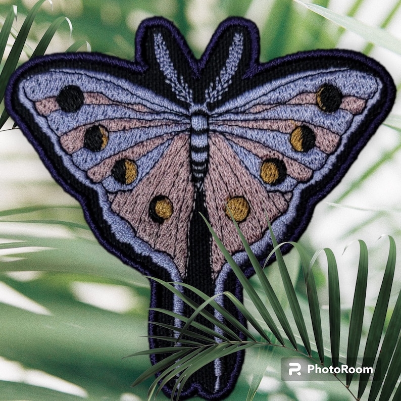 Op de afbeelding: Geborduurde vlinderpatch met een gedetailleerd ontwerp. De patch heeft een kleurenpalet van lavendel, roze en goud, met een zwarte omtrek. De vleugels van de vlinder hebben een gelaagd patroon en cirkelvormige accenten. De patch is geplaatst tegen een vage groene achtergrond.