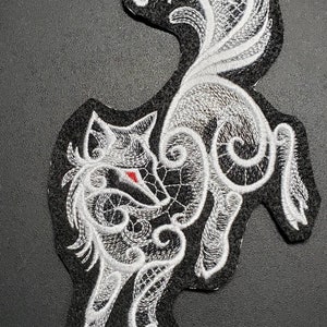 May include: White embroidered patch of a wolf with a red eye, with intricate details and a black background. The wolf is in a curled position with its tail curled around its body. The patch is medium size.