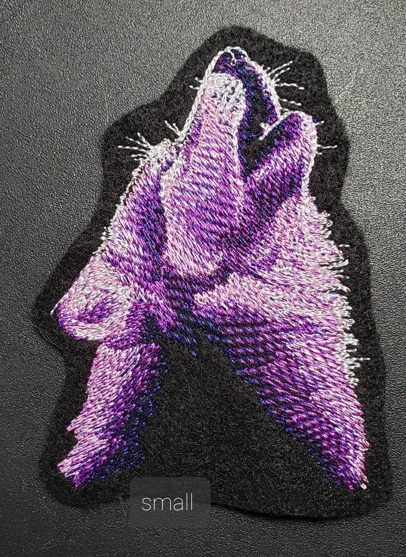 May include: Embroidered patch featuring a howling wolf in shades of purple and white against a black background. The patch has a textured appearance, with the word "small" printed at the bottom. This patch is suitable for jackets, bags, or other fabric items.