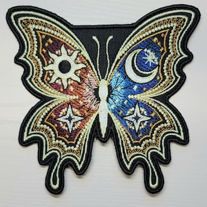 May include: A black embroidered patch of a butterfly with a sun and moon design. The butterfly's wings are decorated with stars and a celestial pattern. The sun is on the left wing and the moon is on the right wing.