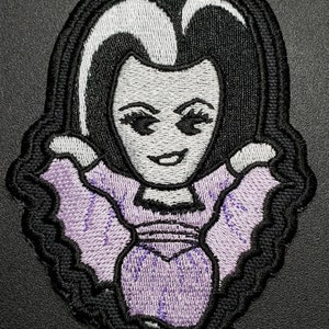 May include: Embroidered iron-on patch featuring a cartoon woman with black hair, white skin, and a purple dress. She has a wide smile and large eyes.