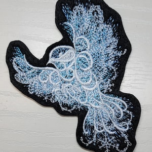 May include: A blue and white embroidered bird patch on a black felt background. The bird is in flight and has a delicate, intricate design.