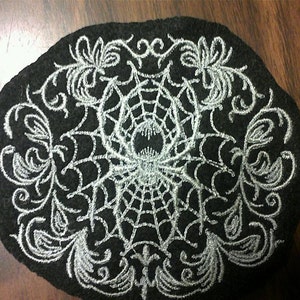 May include: A black felt circle with a white embroidered spider web design. The web is surrounded by intricate floral patterns.