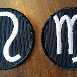 May include: Two black fabric patches with white embroidered zodiac signs. The left patch features the Leo zodiac sign and the right patch features the Virgo zodiac sign.