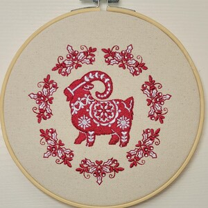 May include: A red and white embroidered goat design is centered on a beige fabric stretched within a wooden embroidery hoop. The goat is surrounded by a wreath of red holly and berries, creating a festive, handcrafted piece.