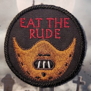 May include: A black and brown embroidered patch with the text "EAT THE RUDE" in red. The patch features a brown mask with white teeth.