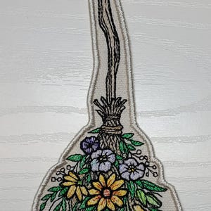 May include: Embroidered patch featuring a broom with a wooden handle and a bundle of colorful flowers. The flowers include yellow, purple, and white blooms with green leaves. The patch has a white border.