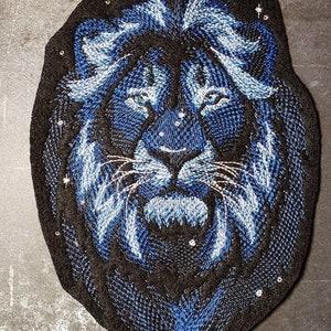 May include: Embroidered patch of a blue lion's face with a black background. The lion's eyes are blue and the mane is detailed with white stitching.