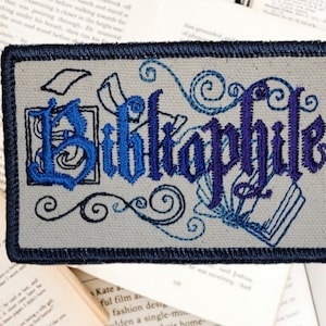 May include: An embroidered patch with the word "Bibliophile" in blue and purple lettering. The patch is surrounded by decorative swirls and images of open books.