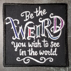 Op de afbeelding: Zwarte geborduurde patch met witte tekst die "Be the weird you wish to see in the world" luidt.