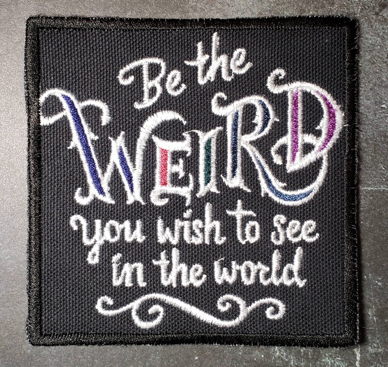 Op de afbeelding: Zwarte geborduurde patch met witte tekst die "Be the weird you wish to see in the world" luidt.