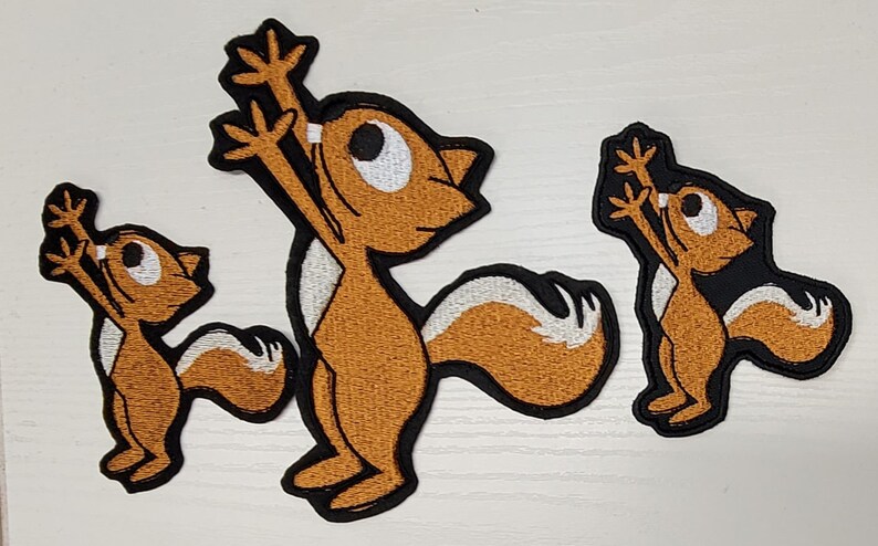 Peut inclure: Trois patchs brod&eacute;s thermocollants repr&eacute;sentant des &eacute;cureuils de dessin anim&eacute; les bras lev&eacute;s en l'air. Les &eacute;cureuils sont bruns avec des accents blancs et des contours noirs.