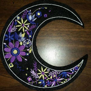 May include: A black crescent moon embroidered with purple, blue, and yellow flowers. The moon is outlined in white thread.