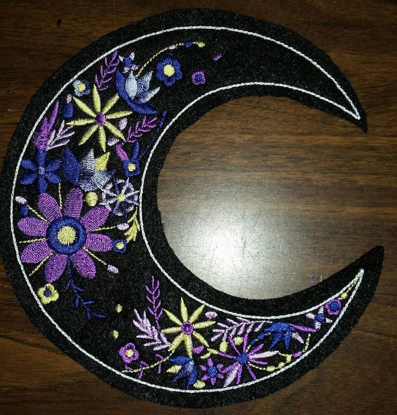 May include: A black crescent moon embroidered with purple, blue, and yellow flowers. The moon is outlined in white thread.
