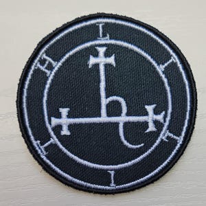 May include: A black embroidered patch with a white sigil. The sigil is a circle with a cross inside, and a letter 'L' at the top and bottom of the circle. The sigil is surrounded by a circle with the letters 'H' and 'I' at the top and bottom.
