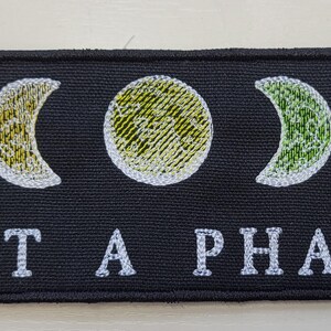 May include: Black rectangular patch featuring embroidered phases of the moon in red, yellow, green, and blue. The text "NOT A PHASE" is embroidered in white below the moon phases. The patch is likely for clothing or accessories.