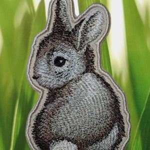 May include: A gray and brown embroidered bunny rabbit patch with a white outline. The patch is on a green background.