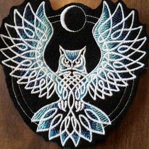 May include: A blue and white embroidered patch of an owl with outstretched wings, surrounded by a crescent moon and a circle. The owl is in a Celtic knot design.