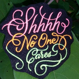 May include: A black embroidered patch with the text "Shhh, No One Cares" in pink, orange, and green thread.