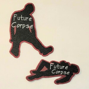 May include: Two black embroidered patches with red outlines. The patches are shaped like silhouettes of people. One patch shows a person walking and the other shows a person lying down. Both patches have the text "Future Corpse" in white.